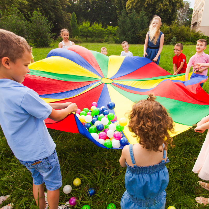 Kinder spielen gemeinsam mit einem bunten Schwungtuch und vielen bunten Bällen auf einer Wiese im Freien.